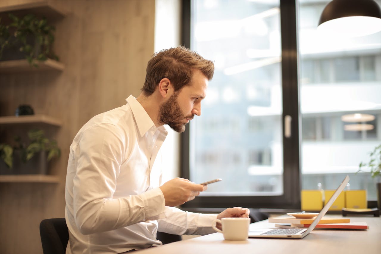Man holding a cell phone while looking at a laptop